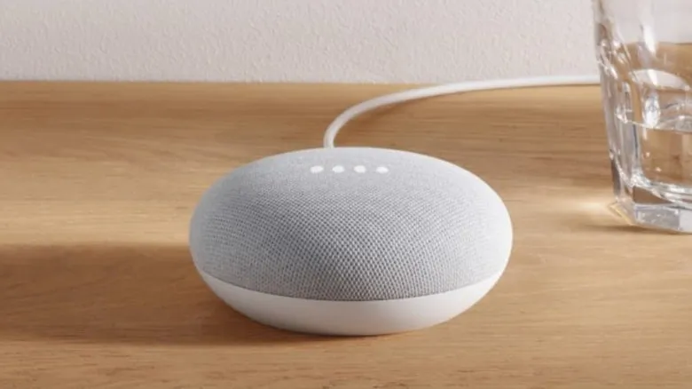 Google Nest Mini speaker in white color on a wooden table next to a glass of water