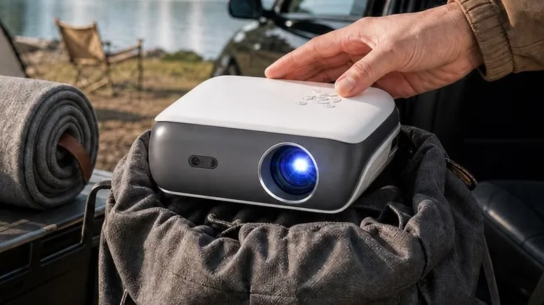 An individual's right hand is on the Happrun Google TV 4K Smart Projector and the device appears to be at a campsite