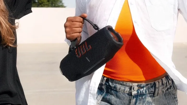 A closeup of an individual dressed in white and orange holding the JBL Charge 6 in their right hand
