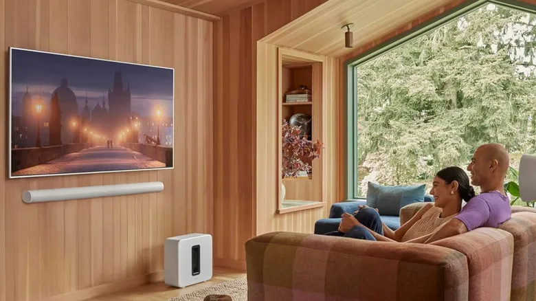 A couple relaxing in a modern living room, watching a wall-mounted TV. A Sonos Arc Ultra soundbar is mounted below it, and a Sonos Sub is placed below the bar.