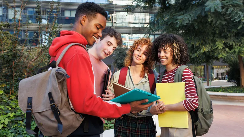 Four college students standing on campus looking at a closed brown book and a closed blue folder