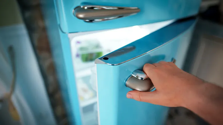 A person's hand seen opening an old refrigerator with latching handles.