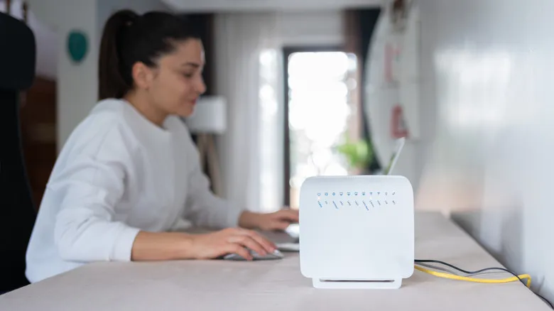 A wireless router sitting atop a desk with someone browsing a laptop in the background.