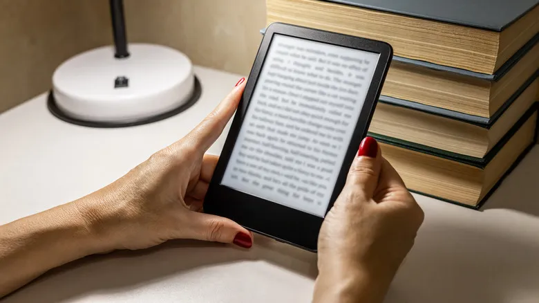 Person reading an ebook on Kindle next to stack of physical books