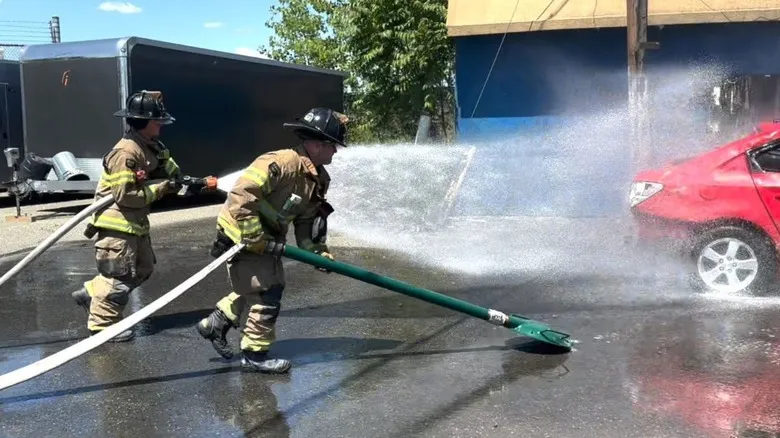 Two firefighters showcasing how to treat car fires. One is spraying water with a hose with the other holds a green pole connected to a turtle-shell looking object on one side and the water supply on the other