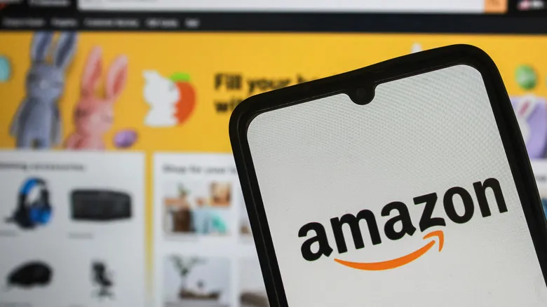 A photograph of the Amazon logo on a smartphone in front of a screen displaying the Amazon homepage.