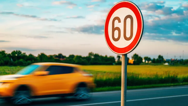 A yellow car races past a red sign that states the country road's speed limit is 60.